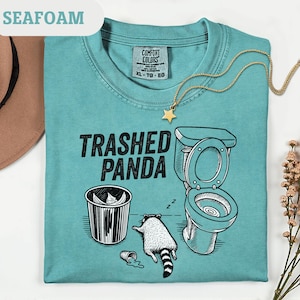 May include: Seafoam green t-shirt featuring the text "Trashed Panda" and a graphic of a raccoon, toilet, and bin. A gold star necklace is draped across the shirt. The shirt is a Comfort Colors XL.