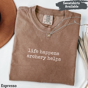 May include: Espresso-colored t-shirt with the text "life happens archery helps" in white. The shirt has a crew neck and short sleeves. A silver necklace with a pendant is draped across the shirt. The words "Sweatshirts Available" are in a banner at the top.
