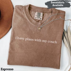 Può includere: T-shirt color espresso con la scritta bianca "i have plans with my couch". La maglietta ha girocollo e maniche corte. Una collana d'argento con un ciondolo è drappeggiata sulla maglietta. Le parole "Sweatshirts Available" sono in alto a destra.