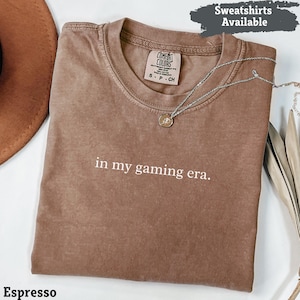 Può includere: T-shirt color espresso con la scritta bianca "in my gaming era". La maglietta ha un girocollo e maniche corte. Una collana d'argento con un ciondolo è drappeggiata sulla maglietta. Le parole "Sweatshirts Available" sono in uno striscione in alto.