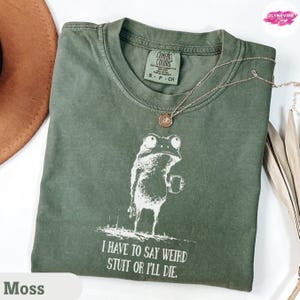 Può includere: Una maglietta verde muschio con una grafica bianca di una rana che tiene una tazza. Il testo "I HAVE TO SAY WEIRD STUFF OR I'LL DIE" è sotto. Una collana d'oro è drappeggiata sulla maglietta. La maglietta ha un'etichetta Comfort Colors.