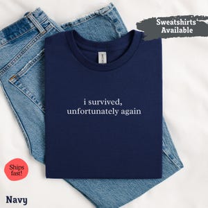 May include: A navy blue t-shirt with the white text "i survived, unfortunately again". The t-shirt is folded on top of blue denim jeans. The words "Sweatshirts Available" are in a grey box in the upper right corner.