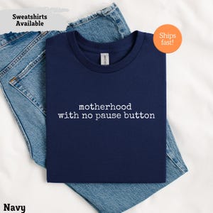 Può includere: T-shirt blu navy con la scritta bianca "motherhood with no pause button". La maglietta è piegata sopra dei jeans blu. Le parole "Sweatshirts Available" sono in un riquadro grigio chiaro in alto a sinistra.