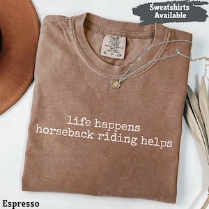 Könnte beinhalten: Espressofarbenes T-Shirt mit dem weißen Text "life happens horseback riding helps". Das Shirt hat einen Rundhalsausschnitt und kurze Ärmel. Eine silberne Halskette mit Anhänger liegt über dem Shirt. Der Text "Sweatshirts Available" steht oben rechts.