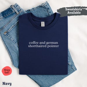 Può includere: T-shirt blu navy con la scritta bianca "coffee and german shorthaired pointer". La maglietta è piegata sopra un paio di jeans blu. Le parole "Sweatshirts Available" sono in un riquadro grigio in alto a destra.