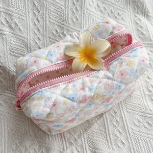 Quilted cosmetic bag with pastel floral print, pink zip, and yellow flower placed on top, shown on a white textured surface.