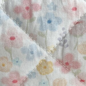 May include: Close-up of a quilted fabric with a floral pattern. The fabric features a white background with scattered flowers in pastel shades of pink, blue, yellow, and peach. The quilting is done with white thread in a diamond pattern.