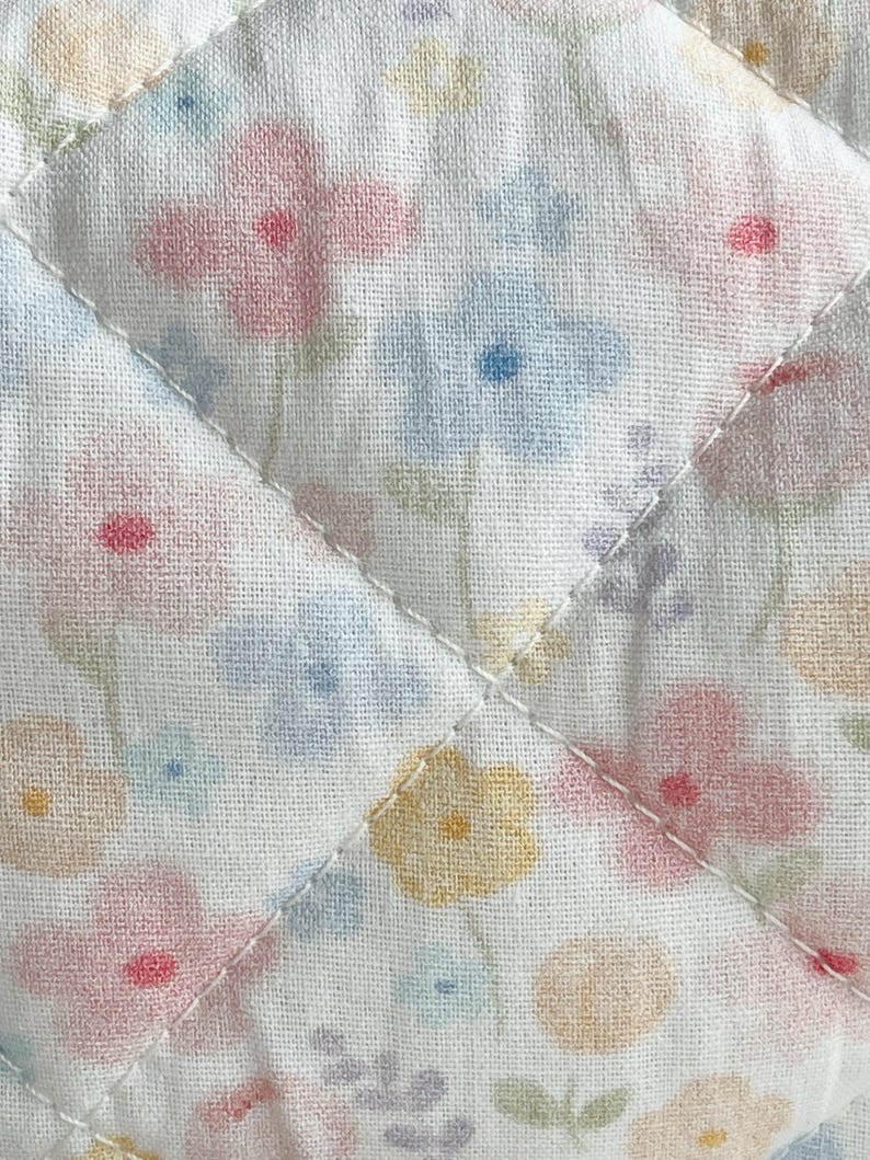 May include: Close-up of a quilted fabric with a floral pattern. The fabric features a white background with scattered flowers in pastel shades of pink, blue, yellow, and peach. The quilting is done with white thread in a diamond pattern.