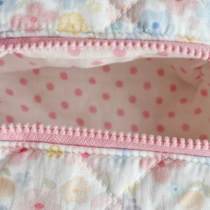 May include: Close-up of a quilted fabric with a pink zipper. The fabric has a floral pattern in pastel colors. The interior lining is white with pink polka dots. The zipper is a light pink color.