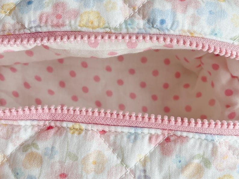 May include: Close-up of a quilted fabric with a pink zipper. The fabric has a floral pattern in pastel colors. The interior lining is white with pink polka dots. The zipper is a light pink color.