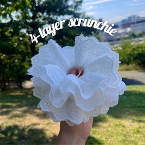 May include: A white, 4-layer scrunchie made of lace fabric. The scrunchie is held up against a background of green grass and trees. The text "4-layer scrunchie" is written in white cursive across the top of the image.