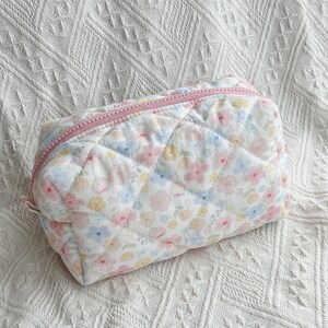 May include: A quilted cosmetic bag with a floral pattern in pastel pink, blue, and yellow. The bag has a pink zipper and a rectangular shape. The bag is on a white textured surface.
