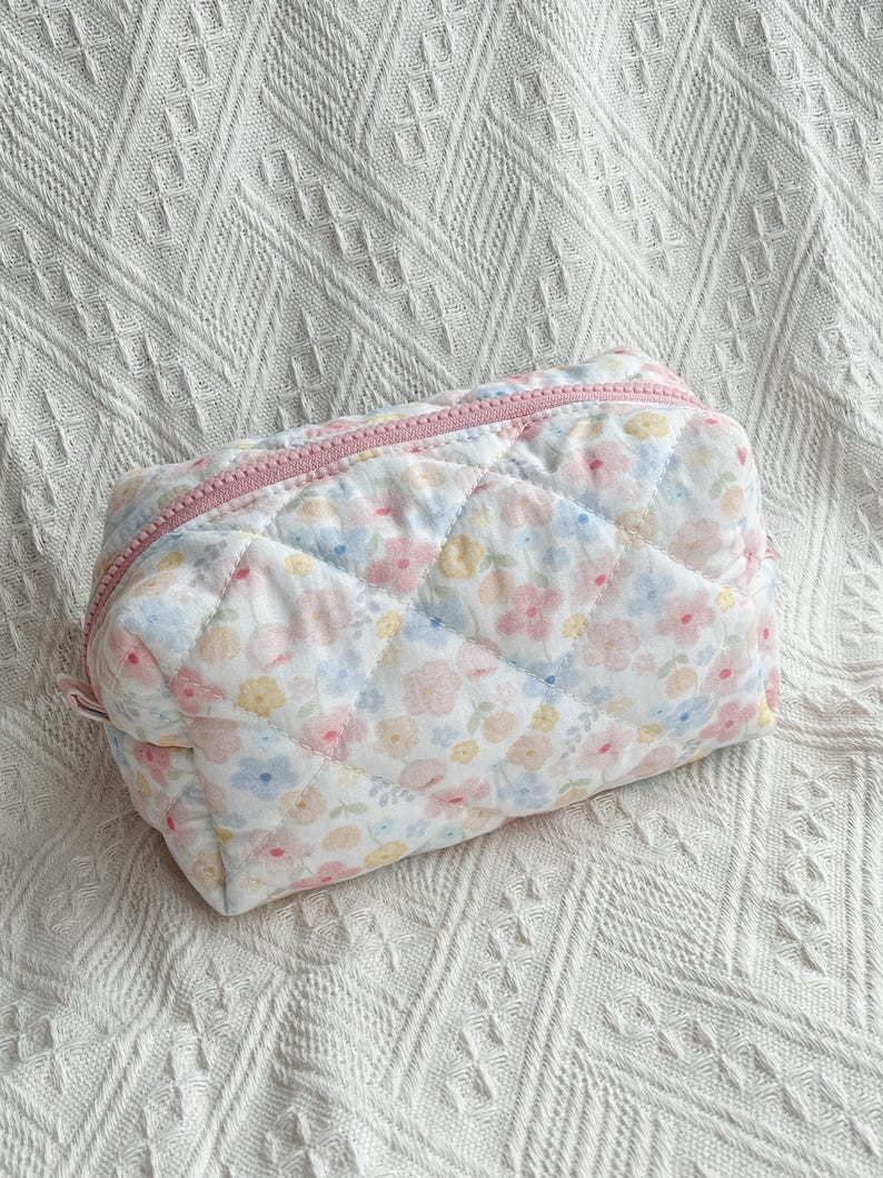 May include: A quilted cosmetic bag with a floral pattern in pastel pink, blue, and yellow. The bag has a pink zipper and a rectangular shape. The bag is on a white textured surface.