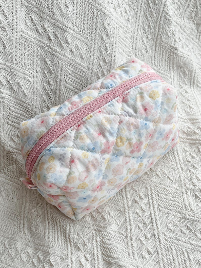 Quilted cosmetic bag with pastel floral print, pink zip, diamond-stitched texture, and rectangular shape on a white surface.