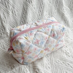 May include: A quilted cosmetic bag with a floral pattern in pastel colors. The bag has a pink zipper and a rectangular shape. The fabric has a diamond pattern.