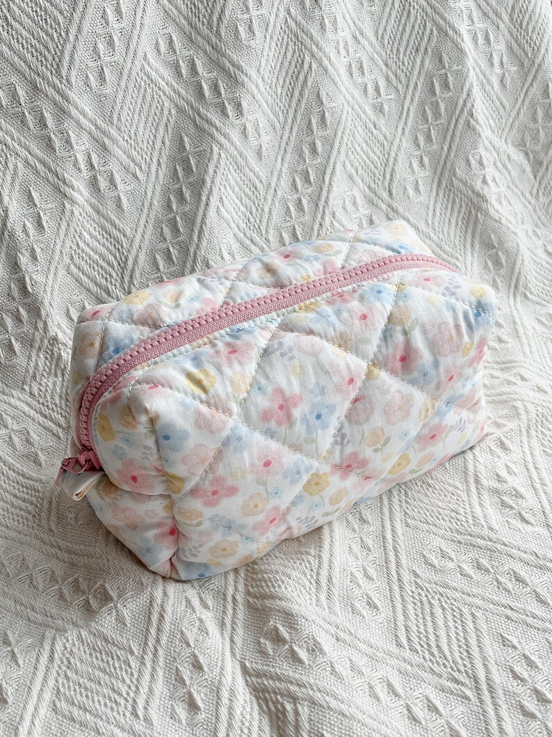 May include: A quilted cosmetic bag with a floral pattern in pastel colors. The bag has a pink zipper and a rectangular shape. The fabric has a diamond pattern.