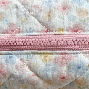 May include: Close-up of a quilted fabric with a floral pattern in pastel colors. A pink zipper runs horizontally across the center. The fabric is white with a repeating pattern of pink, yellow, and blue flowers. The quilting creates a diamond pattern.