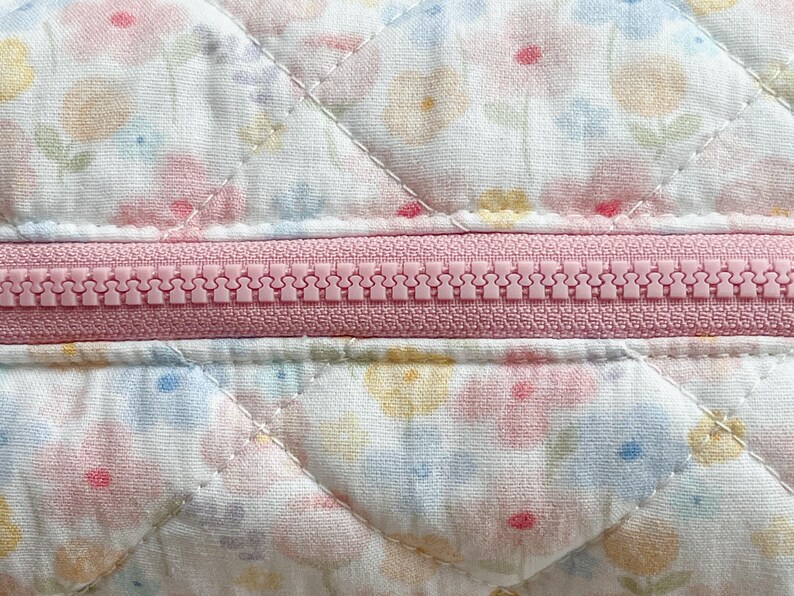May include: Close-up of a quilted fabric with a floral pattern in pastel colors. A pink zipper runs horizontally across the center. The fabric is white with a repeating pattern of pink, yellow, and blue flowers. The quilting creates a diamond pattern.