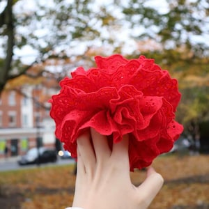 Peut inclure: Une fleur rouge vif et froissée en tissu ajouré. La fleur est tenue devant un arrière-plan extérieur flou d'arbres et de bâtiments. Les détails complexes et la couleur vive de la fleur en font un élément visuel saisissant.