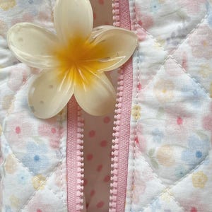 May include: Close-up of a white and yellow flower-shaped object resting on a pink zipper. The zipper is on a quilted fabric with a floral pattern in pastel colors. The interior fabric has a pink polka dot design.