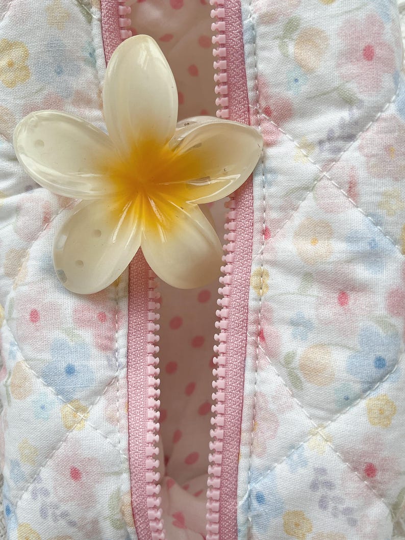 May include: Close-up of a white and yellow flower-shaped object resting on a pink zipper. The zipper is on a quilted fabric with a floral pattern in pastel colors. The interior fabric has a pink polka dot design.