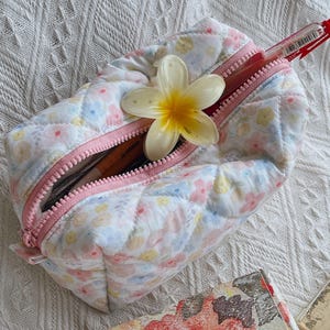May include: A quilted floral print pencil case with a pink zipper, open to reveal pens and pencils. A white and yellow flower rests on top. The case is on a white textured surface.