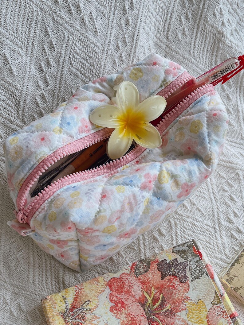 May include: A quilted floral print pencil case with a pink zipper, open to reveal pens and pencils. A white and yellow flower rests on top. The case is on a white textured surface.