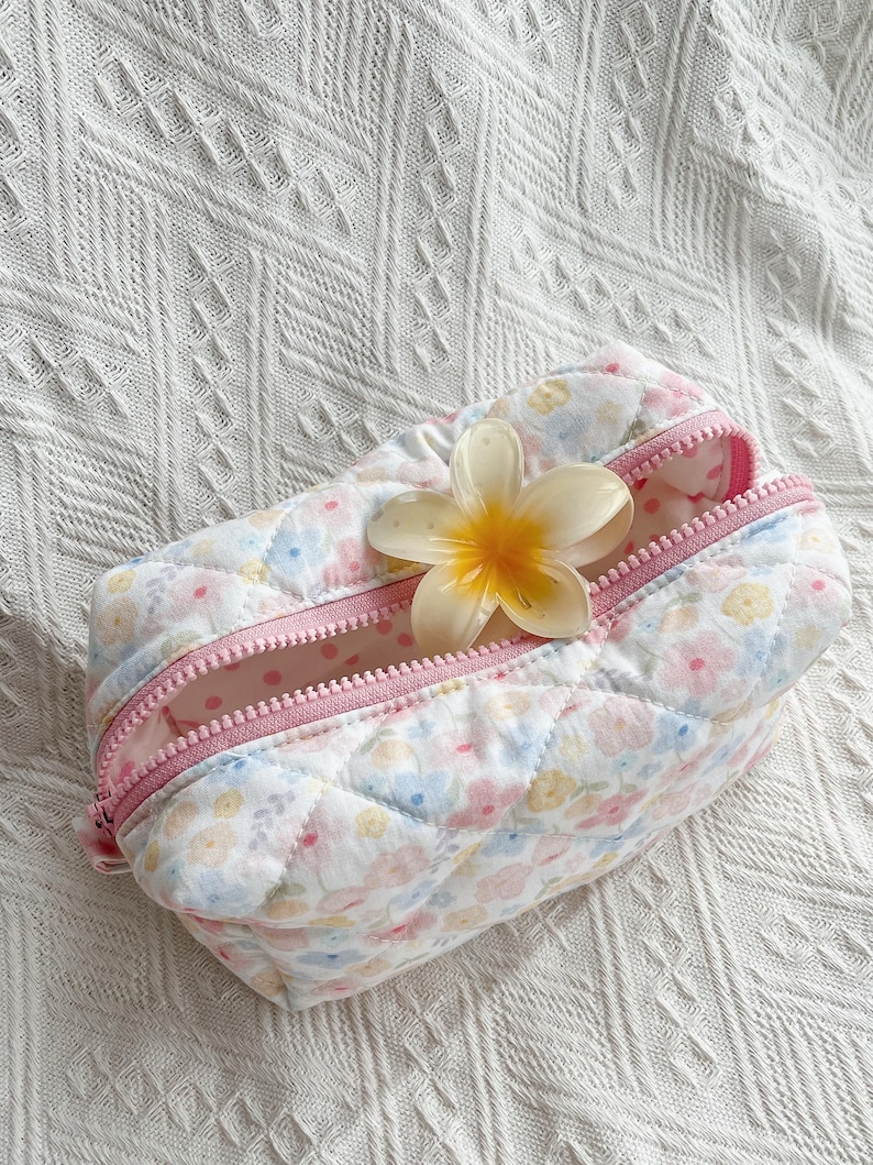 Quilted cosmetic bag with pastel floral print, pink zip, and yellow flower placed on top, shown on a white textured surface.