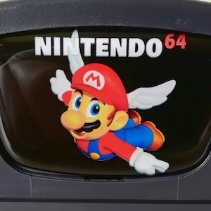 Può includere: Una console Nintendo 64 nera con una placca frontale in plastica trasparente che presenta un'immagine colorata di Mario con le ali. Il testo "NINTENDO 64" è stampato in bianco sulla placca frontale.