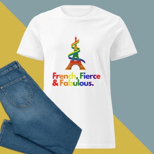 Könnte beinhalten: Weißes T-Shirt mit einem farbenfrohen Grafikdesign des Eiffelturms und einem Regenbogenband. Der Text "French, Fierce & Fabulous" ist darunter gedruckt. Blaue Jeans sind sichtbar.