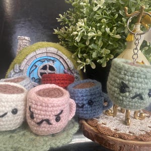 Könnte beinhalten: Gehäkelte Kaffeetassen-Schlüsselanhänger in Weiß, Rosa und Blau, jeweils mit einem einzigartigen Gesichtsausdruck. Eine grüne gehäkelte Tasse mit einem wütenden Gesicht hängt an einem Schlüsselanhänger. Die Szene umfasst ein Miniatur-Hobbit-Haus und Grünzeug.