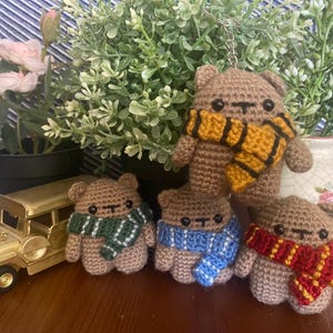 Könnte beinhalten: Gehäkelte braune Bärenfiguren mit bunten Schals. Die Schals sind grün-weiß, blau-weiß, rot-gold und gelb-schwarz. Ein Bär ist ein Schlüsselanhänger. Die Bären werden mit einem goldenen Spielzeugauto und Topfpflanzen präsentiert.