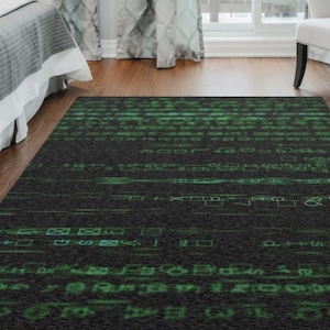 Puede incluir: Una alfombra rectangular con un fondo gris oscuro y un patrón verde estilo matriz. El diseño presenta líneas horizontales de caracteres y símbolos verdes, creando una estética digital. La alfombra está colocada sobre un suelo de madera.