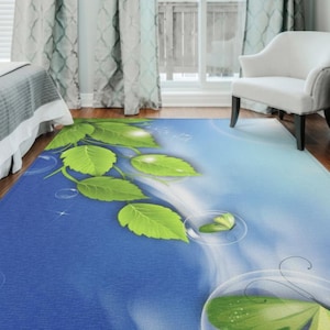 May include: A rectangular area rug with a blue and white gradient background, featuring large green leaves and water droplet designs. The rug is placed on a hardwood floor, with a white chair and curtains visible in the background.