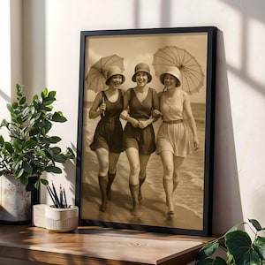 May include: Sepia-toned framed print showing three women in vintage swimwear walking on a beach, each with an umbrella. The women wear hats and swimsuits, one in a dress. The print is in a black frame, displayed on a wooden surface.