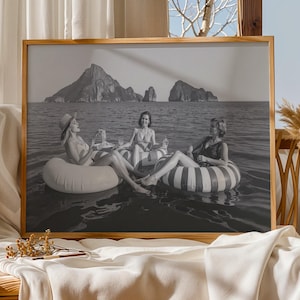 Könnte beinhalten: Schwarz-Weiß-Druck in einem Holzrahmen, der drei Personen in Schwimmreifen im Meer zeigt. Im Hintergrund sind Berge zu sehen. Der Druck steht auf einem Holztisch mit Dekoration.