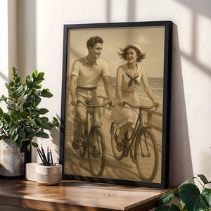 Könnte beinhalten: Gerahmter Sepia-Druck eines Paares, das auf einer Strandpromenade Fahrrad fährt. Der schwarze Rahmen umgibt das Kunstwerk im Vintage-Stil, das einen lächelnden Mann und eine Frau auf Fahrrädern zeigt. Der Druck wird auf einer Holzoberfläche präsentiert.