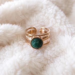 Peut inclure: Une bague dorée ornée d'une pierre centrale circulaire vert foncé à la texture granuleuse. L'anneau est composé de trois fils métalliques fins et parallèles qui se rejoignent sur les côtés, formant un design ouvert et ajustable.