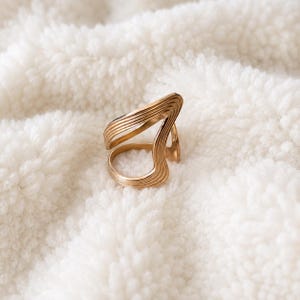 Op de afbeelding: Een goudkleurige metalen ring met een open, asymmetrisch ontwerp en meerdere parallelle gegroefde lijnen langs het oppervlak van de band. De ring heeft een brede, gebogen vorm die uitloopt in een cirkelvormige opening voor de vinger.