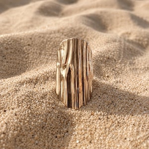 May include: A gold-toned ring featuring a tall, vertical, and curved surface with a highly reflective, irregular, and rippled texture that mimics the appearance of flowing liquid or organic bark patterns, standing upright in a granular sandy texture.