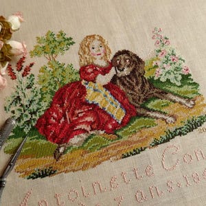 Może przedstawiać: Haft krzyżykowy przedstawiający dziewczynkę w czerwonej sukience z lwem, otoczoną zielenią i kwiatami. Tekst "Antoinette Condé de 7 ans 1851" jest wyszyty różową nicią. Nożyczki i etui na igły znajdują się na pierwszym planie.