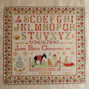 Peut inclure: Une broderie au point de croix vintage sur fond crème. Elle présente un alphabet, des chiffres et des éléments décoratifs rouges, verts, jaunes et bleus. Le texte comprend un nom, un lieu et l'année 1906. Une bordure encadre le motif.