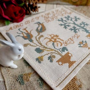 Peut inclure: Un échantillon de point de croix carré, blanc cassé, avec des motifs floraux et d'arbres dans des tons de bleu, marron et jaune. Une petite figurine de lapin en céramique blanche est à gauche. L'échantillon comporte des lettres et des chiffres brodés.