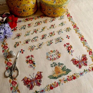 May include: A vintage cross-stitch sampler with floral and butterfly motifs, featuring an alphabet and the year 1815. The sampler is framed by a floral border and includes a thimble and scissors. Two yellow tins are in the background.