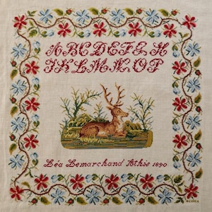 May include: Cross-stitch sampler with a central image of a deer resting on a log, surrounded by greenery. The top border features the alphabet in red, with a floral border of red and blue flowers. The text "Léa Lemarchand Athis 1890" is below the deer.