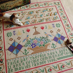 May include: A detailed cross-stitch sampler with floral and geometric patterns in various colors. The sampler features a central floral basket design, a cross, and text in French. A ceramic dog figurine and sewing tools are placed on the sampler.