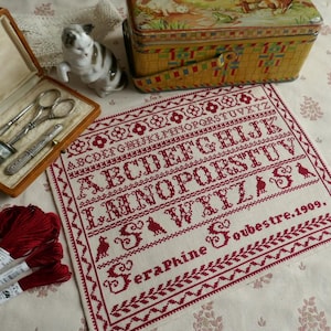 May include: A vintage cross-stitch sampler with red thread on cream fabric, featuring the alphabet and the name "Seraphine Soubestre.1909." The image includes a sewing kit, scissors, and a decorative tin.