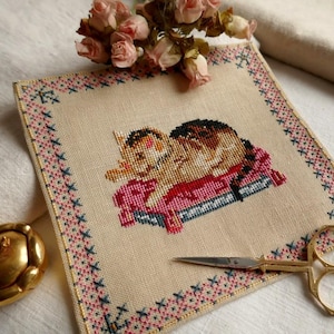 Könnte beinhalten: Kreuzstich-Stickerei mit einer schlafenden Katze auf einem rosa Kissen. Die Katze ist braun und schwarz, und das Kissen befindet sich auf einer blauen Basis. Die Stickerei befindet sich auf einem beigefarbenen Stoff mit einem dekorativen Rand aus rosa und blauen Stichen. Eine goldene Schere und ein goldfarbenes Objekt sind ebenfalls vorhanden.