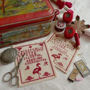 May include: A vintage sewing kit with a red tin box, cross-stitch samplers, red thread spools, and small scissors. The samplers feature red embroidered letters, numbers, and a flamingo design. A small silver compact and other sewing notions are also present.