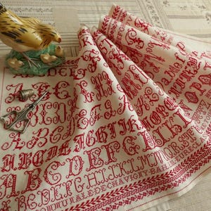 Puede incluir: Tela de lino color crema con letras del alfabeto bordadas en rojo a punto de cruz y borde decorativo. Una figura de gallina de cerámica y herramientas de costura están sobre la tela. La tela está doblada.